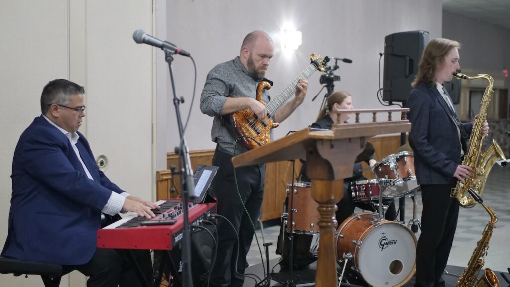 A band performs at an event with members playing keyboards, electric bass, drums, and saxophone.