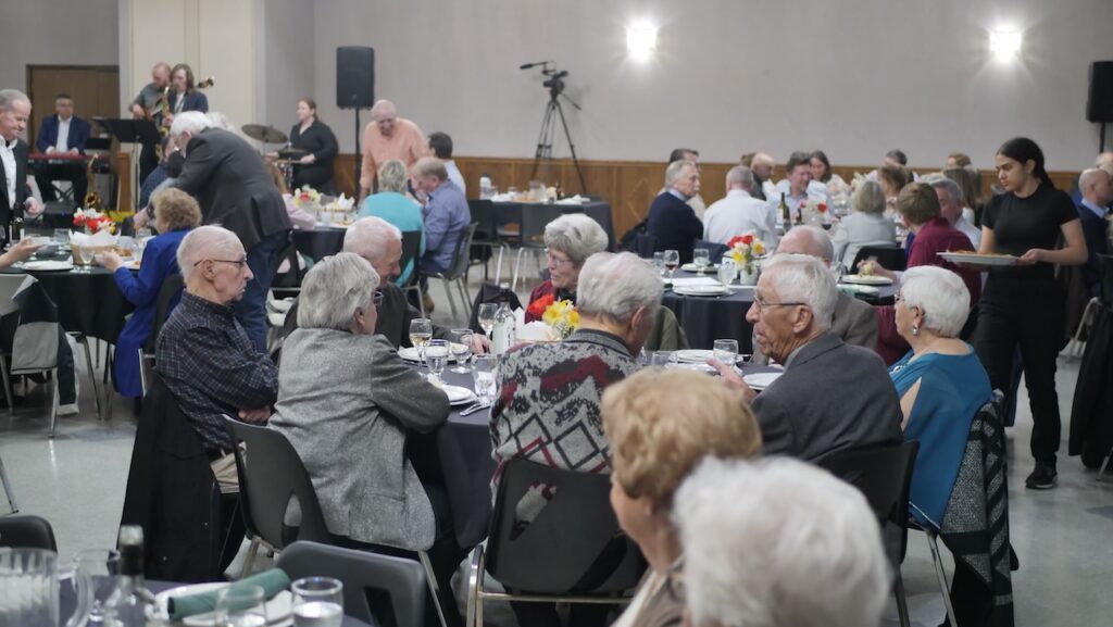 Elderly attendees at a banquet hall with tables set for dining; servers are distributing meals while a presentation is ongoing in the background.