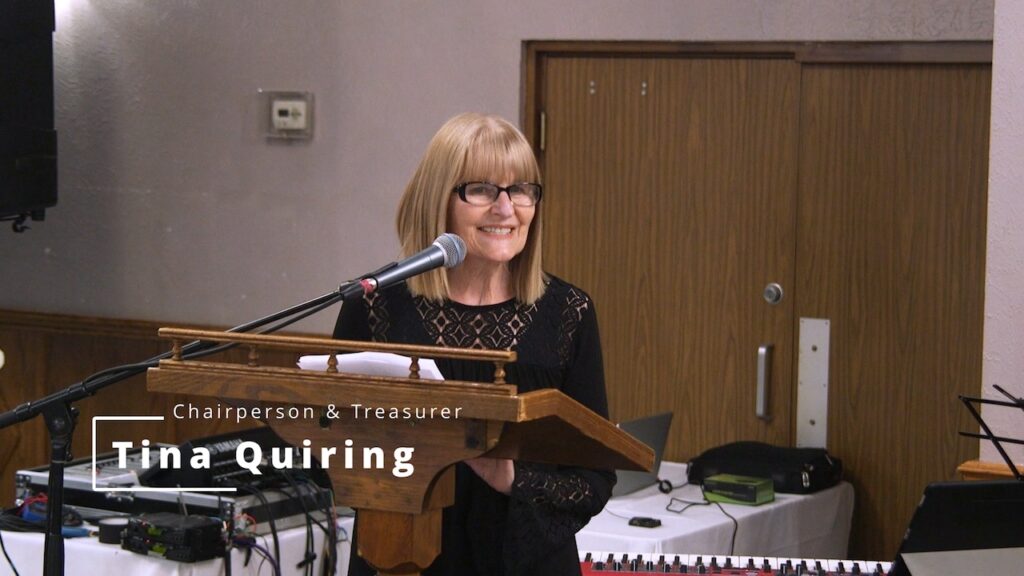 A woman with glasses and a black dress stands at a podium, smiling, with a nameplate reading "chairperson & treasurer tina quiring" in a room with a piano.